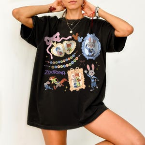 May include: Black t-shirt with colourful Zootopia cartoon graphics. The design includes framed images of characters, a pink bow, and the word "Zootopia" in purple. The shirt has a relaxed fit.