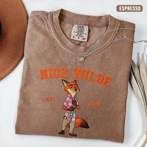 Puede incluir: Camiseta color espresso con el texto "NICK WILDE" bordado en naranja sobre un personaje de zorro de dibujos animados. El zorro lleva una camisa floral rosa y pantalones color caqui. Un collar plateado con un colgante est&aacute; sobre la camiseta.