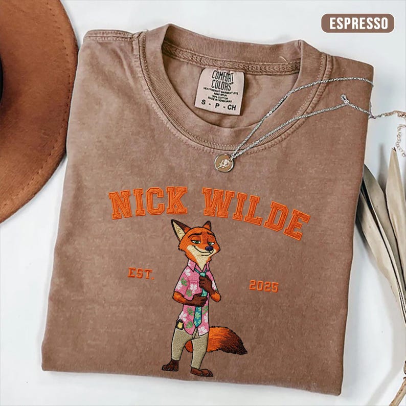 Puede incluir: Camiseta color espresso con el texto "NICK WILDE" bordado en naranja sobre un personaje de zorro de dibujos animados. El zorro lleva una camisa floral rosa y pantalones color caqui. Un collar plateado con un colgante est&aacute; sobre la camiseta.