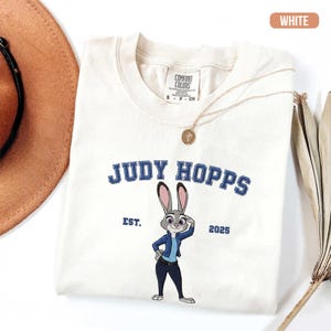 Puede incluir: Camiseta blanca crema con un dibujo animado de Judy Hopps. El dise&ntilde;o incluye el texto "JUDY HOPPS" en azul, y "EST. 2025". Un collar dorado y un sombrero marr&oacute;n tambi&eacute;n son visibles.