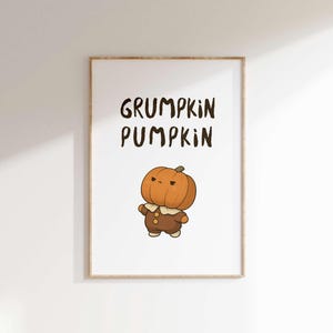 Puede incluir: Impresión enmarcada con un personaje de calabaza de dibujos animados con expresión de enfado. El texto "GRUMPKIN PUMPKIN" está escrito encima de la ilustración. La obra de arte tiene un marco de madera marrón claro y está sobre un fondo blanco.