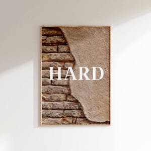 Puede incluir: Una impresión enmarcada de una pared de ladrillo con yeso descascarado. La palabra "HARD" está impresa en letras blancas sobre el yeso.