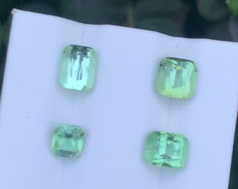 May include: Four light green gemstones with a rectangular cushion cut are displayed on a white surface. The gems vary slightly in size and shade, with a vibrant, translucent appearance. The gems are likely for jewelry making.