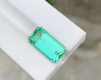 May include: A rectangular, faceted, bright green gemstone. The gem is set against a white surface, with the facets catching the light. The gemstone's color is a vibrant, clear green.