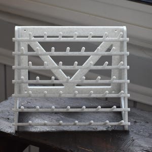 Peut inclure: Un porte-objets mural blanc avec plusieurs rangées de petites chevilles, conçu pour suspendre des articles. Le support a un design géométrique avec une forme en V centrale et des barres horizontales. Il est fait d'un matériau texturé.