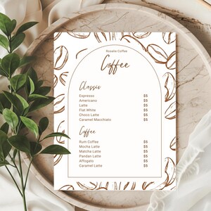May include: A coffee menu from Rosalie Coffee, listing classic and specialty coffee drinks. The white menu has brown text and coffee bean illustrations. It sits on a marble tray, with green leaves in the background.