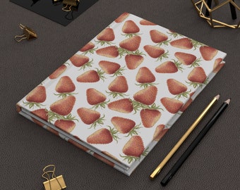 May include: A white notebook with a red strawberry pattern. The notebook is closed and lying on a brown surface with two pencils and a gold metal stand.