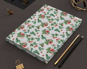 May include: A white notebook with a red and green strawberry pattern. The notebook is closed and lying on a dark gray surface with two pencils and gold paper clips.