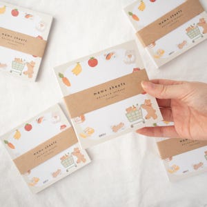 以下が含まれることがあります： クマが食料品でいっぱいのショッピングカートを押しているかわいいデザインのメモ用紙のセット。メモ用紙は白地に茶色の縁取りで、前面には「memo sheets」の文字が印刷されています。クマは茶色で、食料品でいっぱいのショッピングカートを押しています。