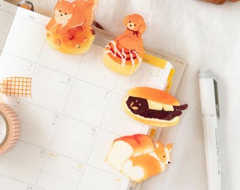 May include: Four clear stickers featuring cartoon animals on top of various pastries. The stickers are on a white calendar with the numbers 7, 8, 9, 14, 15, 16, 22, 23, 24, 29, and 30 printed on it. The stickers feature a brown dog on a donut, a brown dog on a pastry with a swirl of icing, a brown dog on a pastry with a swirl of icing, and a brown dog on a pastry with a swirl of icing.