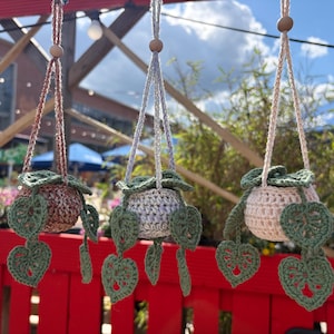 Peut inclure: Trois cache-pots suspendus en crochet. Chacun a un pot rond avec un motif de feuilles vertes et des feuilles vertes suspendues. Les pots sont dans des tons de marron, de gris et de cr&egrave;me. Les cintres sont en cordon torsad&eacute; et ont des perles en bois.