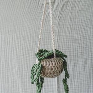 Peut inclure: Un pot de fleurs suspendu en crochet beige fait &agrave; la main, avec une perle en bois et une corde blanc cass&eacute;. Le pot contient des feuilles en crochet vert, cr&eacute;ant un aspect d&eacute;coratif et naturel. Parfait pour les petites plantes.