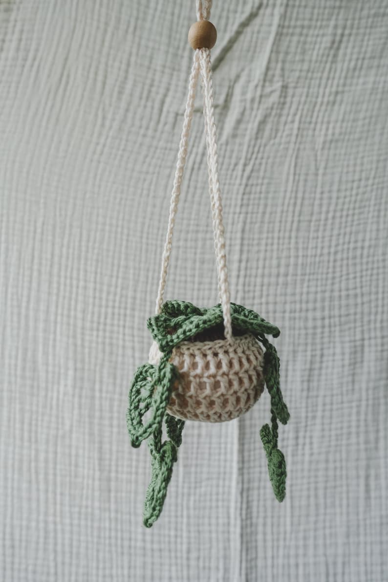 Peut inclure: Un pot de fleurs suspendu en crochet beige fait &agrave; la main, avec une perle en bois et une corde blanc cass&eacute;. Le pot contient des feuilles en crochet vert, cr&eacute;ant un aspect d&eacute;coratif et naturel. Parfait pour les petites plantes.