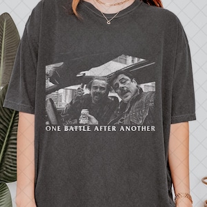 May include: Dark gray t-shirt featuring a black and white image of two men in a car. The image includes the text "SMALL ONE BATTLE AFTER ANOTHER" in white font. The shirt is a casual, oversized style.