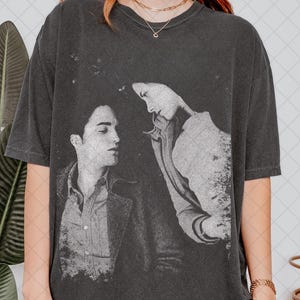 May include: Dark gray t-shirt featuring a black and white image of two people. The image appears to be a still from a movie or television show. The t-shirt has a crew neck and short sleeves.