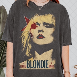Może przedstawiać: Ciemnoszara koszulka z grafiką vintage Blondie z Debbie Harry. Projekt zawiera czerwoną gwiazdę, nazwę zespołu i "New York City 1977" w stylu retro. Koszulka ma klasyczny okrągły dekolt.