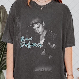 May include: Dark grey t-shirt with a black and white image of Mac DeMarco wearing a hat. The text "Mac DeMarco" is written in teal, stylized font across the front. The shirt has short sleeves and a relaxed fit.