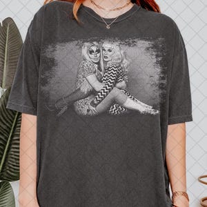 Puede incluir: Camiseta gris oscuro con un gráfico en blanco y negro de dos personas abrazándose. El gráfico tiene un aspecto vintage y desgastado. La camiseta tiene un corte holgado, mangas cortas y cuello redondo. El estilo general es casual y cómodo.