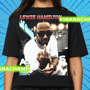 Może przedstawiać: Czarny t-shirt z grafiką Lewisa Hamiltona w okularach przeciwsłonecznych i łańcuchem, pokazującego środkowy palec. Tekst "LEWIS HAMILTON" znajduje się nad obrazem w kolorze czerwonym. Koszulka ma okrągły dekolt.