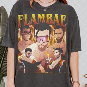 May include: Dark gray t-shirt featuring a graphic design with the word "FLAMBAE" in orange and yellow. The design includes illustrations of several men wearing sunglasses and holding objects, with a retro style.