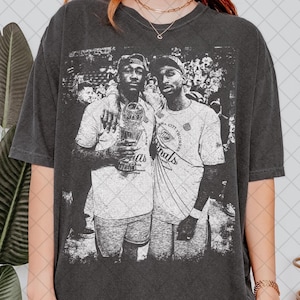 Op de afbeelding: Donkergrijs t-shirt met een zwart-wit afbeelding van twee mannen, waarvan één een trofee vasthoudt. Het shirt heeft een vintage, distressed look. De tekst "Finals" is zichtbaar op het shirt.