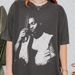 May include: Dark gray t-shirt featuring a black and white graphic of a person holding a guitar. The person is wearing a white sleeveless shirt. The t-shirt has a vintage, worn look.