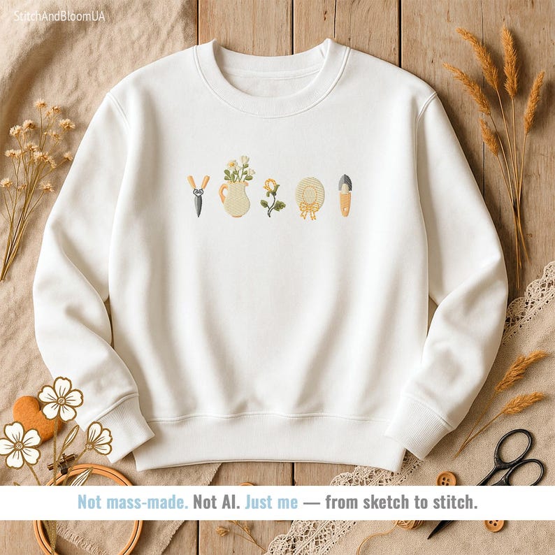 May include: White sweatshirt with embroidered garden-themed designs, including a watering can, flowers, and gardening tools. The text "Not mass-made. Not AI. Just me - from sketch to stitch." is at the bottom.