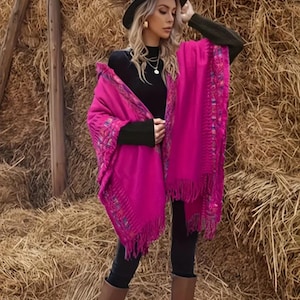 May include: A vibrant fuchsia poncho featuring intricate multicoloured embroidered trim along the edges and long fringe detailing. The garment is worn over a black long-sleeved top and black trousers, paired with brown knee-high boots and a dark fedora hat.