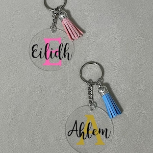 Puede incluir: Dos llaveros de acrílico transparente con un borla rosa y azul. El llavero con la borla rosa tiene el nombre "Eilidh" con una letra E rosa. El llavero con la borla azul tiene el nombre "Ahlem" con una letra A amarilla.