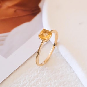 Könnte beinhalten: Ein goldener Ring mit einem quadratischen, gelben Edelstein. Der Ring hat ein zartes Band mit kleinen Steinen um den Edelstein.