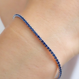 Op de afbeelding: Een delicate tennisarmband met een rij kleine, ronde, blauwe saffieren die in een zilveren band zijn gezet.