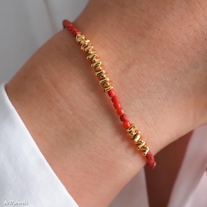 Puede incluir: Una pulsera delicada con pequeñas cuentas rojas intercaladas con cuentas cilíndricas doradas y texturizadas. La pulsera se lleva en la muñeca, mostrando el contraste entre los elementos rojos y dorados. La joya es sencilla y elegante.