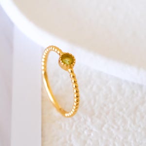 Peut inclure: Une bague en or avec une petite pierre précieuse ronde vert clair. Le bracelet présente un motif de perles. La bague est posée sur un fond blanc, avec un soupçon de motif sur le côté gauche. C'est un bijou.