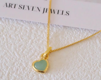 May include: A gold chain necklace with a small heart pendant. The heart is light green and has a gold border. The necklace is on a white surface.