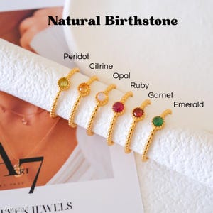 Op de afbeelding: Goudkleurige ringen met natuurlijke geboortestenen, waaronder peridot, citrien, opaal, robijn, granaat en smaragd. De ringen worden getoond op een wit oppervlak met de tekst "Natural Birthstone".