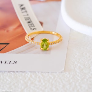 May include: A gold ring featuring a central oval-cut lime green gemstone held by four prongs, with a band accented by a row of small clear stones. The ring is positioned on a white card printed with the text ART7 JEWELS in black lettering.