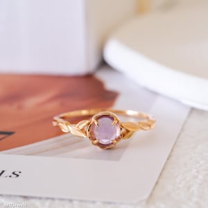 Op de afbeelding: Een gouden ring met een centrale ovaalvormige lichtpaarse edelsteen die door vier pootjes wordt vastgehouden. De band is ontworpen met een reeks verbonden, bladvormige metalen segmenten die uit de zetting komen voor een botanische uitstraling.