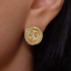 Könnte beinhalten: Goldfarbene Ohrringe mit einem runden Design, das ein stilisiertes Gesicht und die Worte "Lucky Lucky" zeigt, die sich um den Umfang wiederholen.