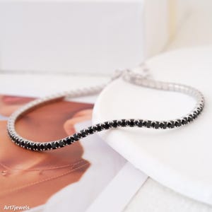 Peut inclure: Un bracelet de tennis argenté orné d'une rangée continue de petites pierres noires. Le bracelet est présenté sur une surface blanche, avec un arrière-plan doux et flou. Les pierres sont espacées uniformément, créant un design élégant.