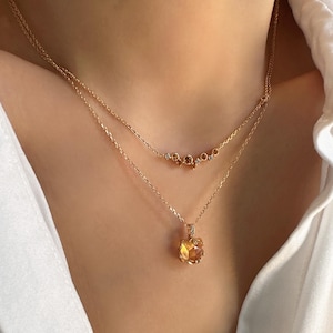 Può includere: Una collana con catena d'oro con un ciondolo a forma di grande pietra preziosa sfaccettata di colore ambrato. Il ciondolo è circondato da pietre preziose più piccole e trasparenti. La collana è sovrapposta a una seconda catena d'oro più sottile con una fila di pietre preziose più piccole e trasparenti.
