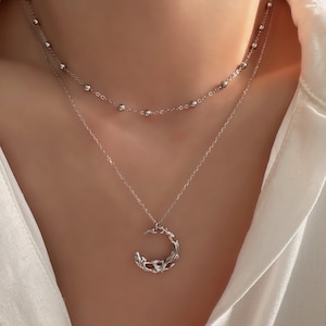 Può includere: Una collana argentata a due fili con una catena delicata e un ciondolo a forma di luna crescente con un design floreale.