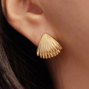 Könnte beinhalten: Nahaufnahme eines goldfarbenen Ohrsteckers in Muschelform. Der Ohrring hat ein fächerartiges Design mit vertikalen Rippen. Der Ohrring wird am Ohr getragen.