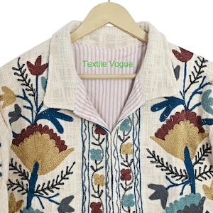 May include: Cream-colored embroidered jacket with floral and leaf patterns in shades of gold, teal, burgundy, and blue. The jacket has a collared neckline and a striped pink and white lining. The text "Textile Vogue" is visible on the inside collar.