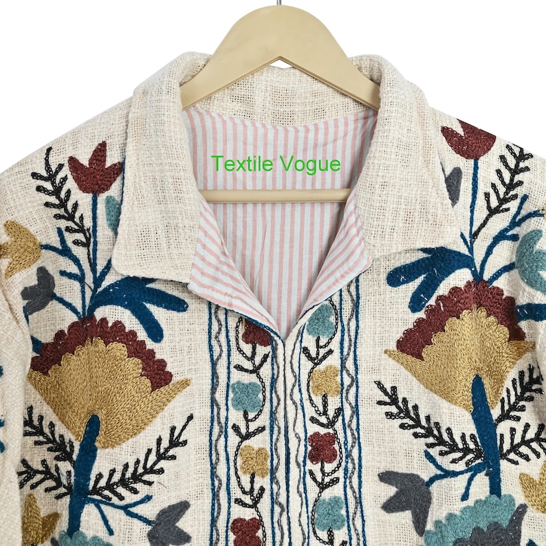 May include: Cream-colored embroidered jacket with floral and leaf patterns in shades of gold, teal, burgundy, and blue. The jacket has a collared neckline and a striped pink and white lining. The text "Textile Vogue" is visible on the inside collar.