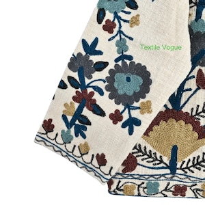 May include: Close-up of a cream-colored textile with intricate floral embroidery in shades of blue, brown, and gold. The design includes stylized flowers and leaves. The text "Textile Vogue" is visible on the fabric.
