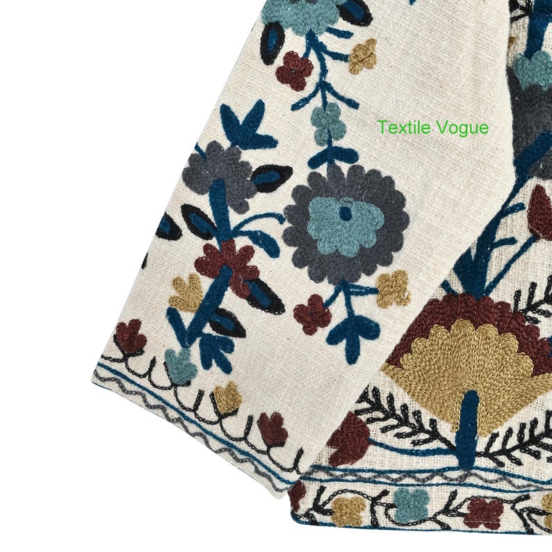 May include: Close-up of a cream-colored textile with intricate floral embroidery in shades of blue, brown, and gold. The design includes stylized flowers and leaves. The text "Textile Vogue" is visible on the fabric.