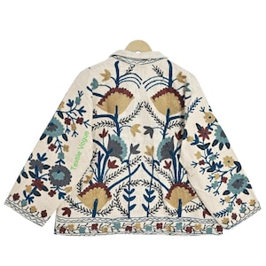 May include: Cream-colored jacket with intricate floral embroidery in shades of blue, brown, and gold. The jacket features a collared neckline and long sleeves, with the text "Textile Vogue" visible on the side.