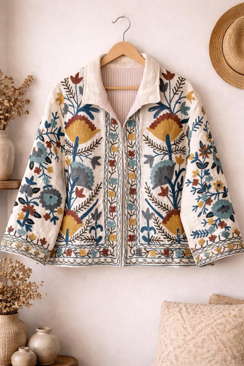 May include: Cream-colored embroidered jacket with a floral design. The jacket features a collar and a zip-up front. The embroidery includes yellow, blue, and brown flowers and leaves. The jacket is hanging on a wooden hanger.
