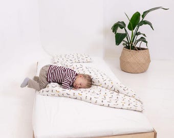 Pode incluir: Uma criança dormindo em uma cama com um colchão branco e uma estrutura de madeira clara. A roupa de cama apresenta um padrão de pequenas formas coloridas. Um travesseiro e um travesseiro longo também são visíveis. Uma planta em vaso está por perto.