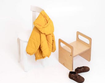 Pode incluir: Uma cadeira de madeira branca com um casaco acolchoado amarelo pendurado. Um pequeno banco de madeira clara com recortes de alça está ao lado da cadeira. Sapatos de camurça castanhos no chão.
